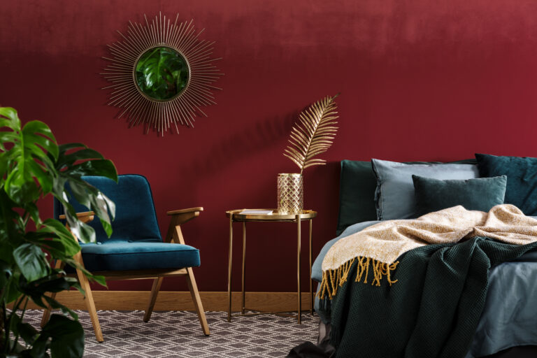 Gold table between green armchair and bed in sophisticated red bedroom interior with mirror